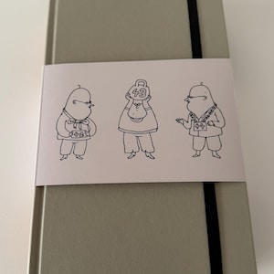 May include: A gray notebook with a black elastic band closure. A white paper band wraps around the notebook, featuring three cartoon figures with the numbers 42, 43, and 44.