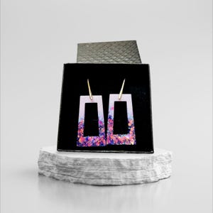 Puede incluir: Un par de pendientes rectangulares con base blanca y purpurina iridiscente rosa, morado y azul. Los pendientes se muestran en una caja negra sobre una superficie blanca.