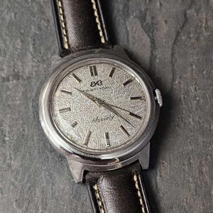 Könnte beinhalten: Eine silberne Armbanduhr mit weißem Zifferblatt und schwarzen Zeigern. Die Uhr hat ein braunes Lederarmband mit weißen Nähten. Das Zifferblatt der Uhr trägt den Text "Eternamatic" und "288".