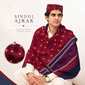 May include: A man wearing a Sindhi Ajrak shawl and cap. The shawl is a deep red with a repeating pattern of white and blue floral designs. The cap has a similar pattern. The text "Sindhi Ajrak" and "Tradition woven in every thread" are visible.