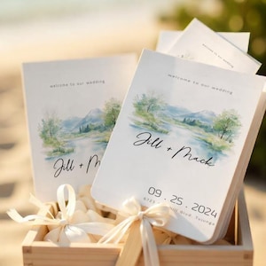May include: Wedding fan favours in a wooden crate. Each fan features a watercolour landscape design with the names "Jill + Mack" and the wedding date. The fans are tied with cream-coloured ribbons.