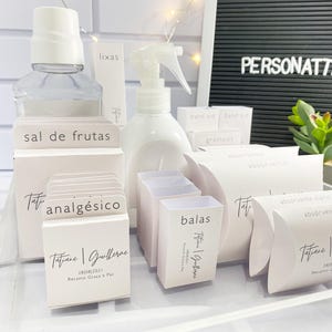 Puede incluir: Una colección de pequeñas cajas blancas con varias etiquetas, incluyendo "sal de frutas", "analgésico" y "balas". Las cajas están dispuestas sobre una superficie blanca y presentan el texto "Tatiane | Guilherme" y una fecha.