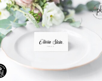 May include: White place card with black calligraphy text that reads "Olivia Stein" and "Table 2".