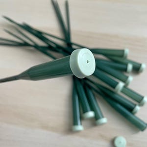 May include: A close-up of a bundle of green plastic flower stems with white caps. The caps have a small hole in the center.