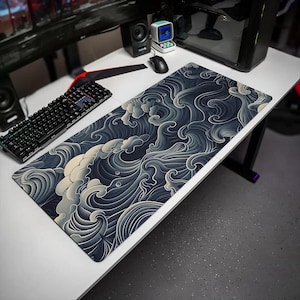 May include: A rectangular desk mat featuring a dark navy blue and cream-colored graphic pattern of stylized, swirling ocean waves. The mat is positioned on a white desk surface next to a black mechanical keyboard, a computer mouse, and a small speaker.