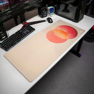 May include: A rectangular desk mat with a light beige surface featuring a graphic design of two overlapping circles in shades of red and orange, accented by thin horizontal lines on the left side and a small logo in the top right corner.