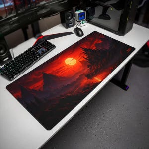 May include: A rectangular desk mat featuring a dark, atmospheric landscape illustration with a vibrant red sun, jagged mountain silhouettes, and a reflective body of water. The design uses a deep red and black color palette across the surface.