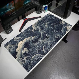 May include: A rectangular desk mat featuring a dark navy blue and cream-colored graphic pattern of stylized, swirling ocean waves. The design covers the entire surface of the mat, which is positioned on a white desk surface with computer equipment.