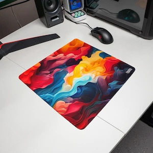 Peut inclure: Un tapis de souris carré présentant un motif abstrait vibrant avec des tourbillons de couleurs rouge, orange, jaune et bleu. La surface affiche une esthétique fluide rappelant de la fumée avec un petit logo blanc imprimé sur le côté droit.