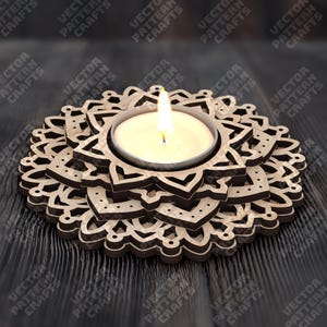 May include: A wooden candle holder with a floral design. The candle holder has a circular shape with layered, laser-cut patterns. A lit tea light candle sits in the center, casting a warm glow. The background is a dark wooden surface.