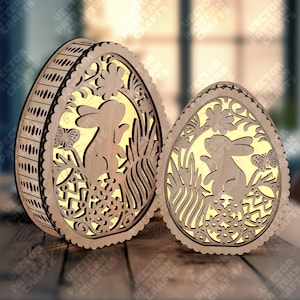 Può includere: Due decorazioni in legno a forma di uovo di Pasqua con intricati disegni intagliati. L'uovo più grande ha una forma cilindrica, mentre quello più piccolo è più piatto. Entrambi presentano un coniglietto, fiori ed erba, con una luce calda che emana dall'interno.