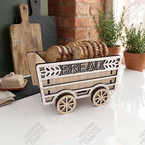 May include: A light-colored wooden bread bin shaped like a wagon with four wheels. The side panel features the word BREAD in dark lettering, flanked by wheat stalk cutouts. The container holds several slices of brown bread inside the open top.
