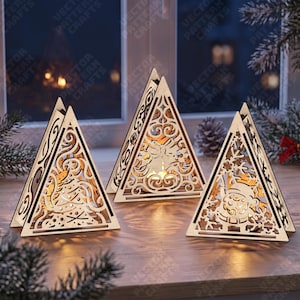 Puede incluir: Tres portavelas triangulares de madera con intrincados diseños cortados con láser. La pieza de la izquierda muestra un muñeco de nieve, la central un motivo de campana y la de la derecha una figura de Papá Noel. Cada marco de madera clara brilla.