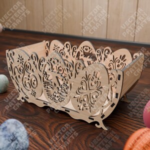 May include: A light-colored wooden basket with intricate laser-cut designs. The basket features an egg-shaped design on the sides and floral patterns. The text "VECTOR PATTERN CRAFTS" is visible on the sides. The basket is on a wooden surface.