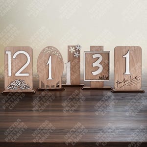 Può includere: Una collezione di segnaposto numerici in legno. Ogni segnaposto presenta un design unico, con numeri, elementi floreali e cornici decorative. I numeri sono bianchi e i segnaposto sono in legno marrone chiaro.