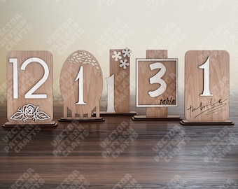 May include: A collection of wooden table number signs. Each sign features a unique design, including numbers, floral accents, and decorative frames. The numbers are in white, and the signs are made of light brown wood.