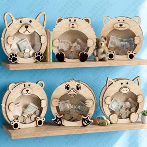 Könnte beinhalten: Sechs runde Spardosen aus Holz mit durchsichtiger Acrylfront, jede in Form eines anderen Tieres wie Hund, Bär oder Schwein. Jede Spardose ist mit einem eingravierten Namen versehen und enthält verschiedene Münzen und Geldscheine.