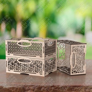 Peut inclure: Deux boîtes décoratives en bois avec des motifs découpés au laser. Une boîte est empilée sur une autre, et une troisième boîte est sur le côté. Les boîtes ont des poignées et sont en bois clair. L'arrière-plan est flou.