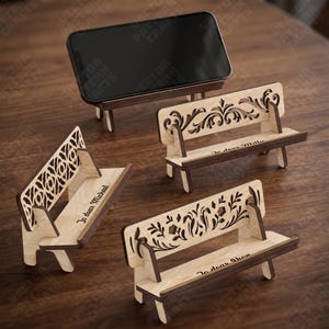 May include: Three small wooden bench-shaped phone stands with intricate laser-cut backrests. One stand holds a black smartphone. The benches feature light wood seats and dark wood edges, with personalized text engraved on the seats reading Jo dear Michael, Jo dear Milly, and Jo dear Shon.