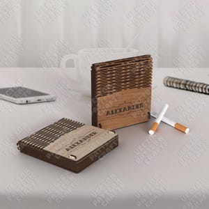 May include: Two wooden cigarette cases, one open and one closed, with the name "ALEXANDER" engraved on them. The cases are light brown with a dark brown trim. Two cigarettes are next to the cases. A white mug and a smartphone are in the background.