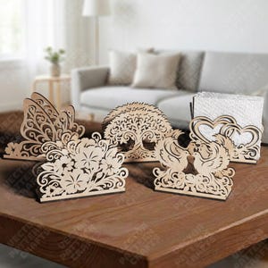 Pode incluir: Uma coleção de porta-guardanapos de madeira cortados a laser com designs intrincados. Os designs incluem uma borboleta, flores, uma árvore, duas pombas e corações. Os porta-guardanapos são exibidos em uma mesa de madeira, mostrando sua natureza decorativa.