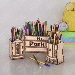 Puede incluir: Soportes de lápices de madera con los nombres "Emily", "Ms. Park" y "Jacob" grabados. Los soportes están llenos de lápices de colores y bolígrafos. Una mochila amarilla y un reloj están en el fondo.
