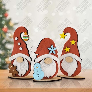 Puede incluir: Tres adornos navideños de gnomos de madera. Cada gnomo tiene un gorro rojo y una barba blanca. Uno sostiene un hombre de jengibre, otro un muñeco de nieve y el tercero estrellas amarillas.