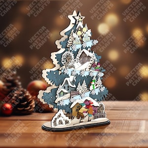 Könnte beinhalten: Weihnachtsbaumschmuck aus Holz mit einer Winterdorf-Szene. Der Baum ist blau und weiß bemalt und zeigt ausgeschnittene Häuser, Bäume und Figuren. Ein Stern ziert die Spitze des Baumes, und Schneeflocken sind verstreut.