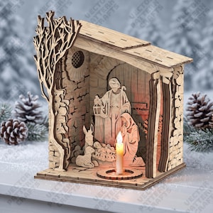May include: A laser-cut wooden nativity scene featuring a stable structure with a tree silhouette on the side. Inside, figures of two adults, a donkey, and a lamb surround a manger. A small battery-operated LED candle sits in the foreground.