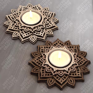 Puede incluir: Dos candelabros de madera con un diseño floral, cada uno con una vela de té encendida. Los candelabros son marrones y tienen intrincados patrones recortados. El fondo es una superficie gris claro.
