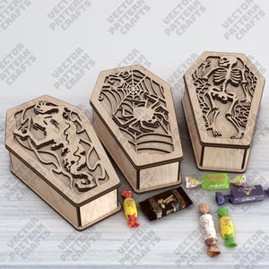 Könnte beinhalten: Drei Sarg-förmige Holzkisten mit Laserschnitt-Designs. Eine zeigt ein Spinnennetz und eine Spinne, eine andere ein Skelett und die dritte eine Kreatur. Verschiedene verpackte Süßigkeiten sind vor den Kisten verstreut. Die Kisten sind aus hellem Holz.