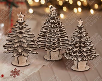 May include: Three wooden Christmas tree decorations. The trees are laser cut with intricate details, featuring star and snowflake toppers. They are displayed on a light-colored surface with blurred background lights.