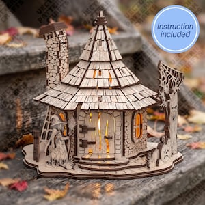 May include: A laser-cut wooden model of a witch's cottage featuring a conical shingled roof, a tall stone-textured chimney, and a central arched door. The structure includes a figure of a witch, a gnarled tree, and a pumpkin. Text reads: Instruction included.