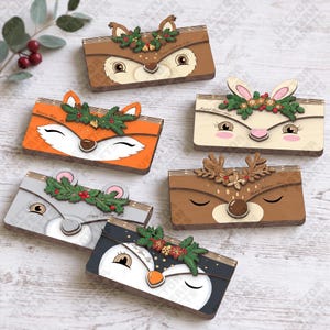 May include: A collection of six rectangular wooden ornaments featuring stylized animal faces including a deer, fox, rabbit, and owl. Each piece is decorated with a festive greenery wreath, berries, and small holiday-themed text details.