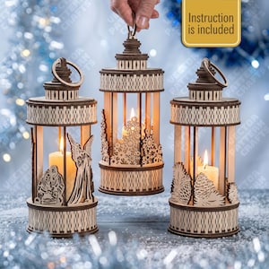 Puede incluir: Tres farolillos cilíndricos de madera con patrones cortados a láser, cada uno con una vela encendida en su interior. Los diseños incluyen un belén con un ángel, copos de nieve y piñas. Un cartel dorado indica Instruction is included.