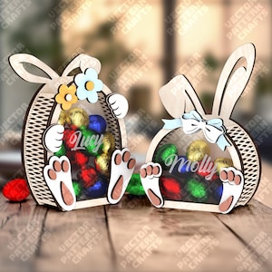 Puede incluir: Dos cestas de madera con forma de conejito de Pascua con frentes transparentes, llenas de caramelos de colores envueltos en papel de aluminio. Una cesta tiene el nombre "Lucy" y flores, la otra "Molly" y un lazo. Las cestas están sobre una superficie de madera.
