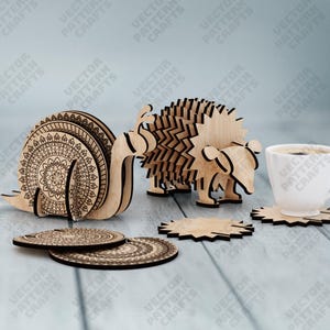 Puede incluir: Posavasos de madera con forma de animales y un soporte a juego. El juego incluye un caracol, un erizo y un oso, junto con varios posavasos redondos. Una taza blanca de caf&eacute; est&aacute; cerca. Los posavasos son de madera clara.
