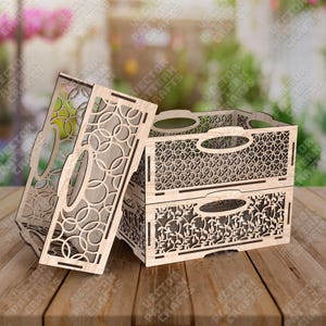 May include: Two-tiered wooden storage crate with decorative cut-out patterns. The top tier features a geometric design, while the bottom tier showcases a floral motif. The crate has handles and is made of light-colored wood. A matching lid is propped up.