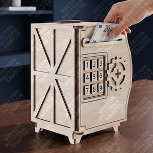 May include: A wooden money bank shaped like a safe, with a combination keypad and a gear design. The bank has a slot for inserting money, and a hand is placing a twenty-euro bill into the slot. The bank is made of light-colored wood.