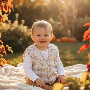 Könnte beinhalten: Ein lächelndes Baby trägt eine cremefarbene, geknöpfte Strickweste mit bunten Sprenkeln über einem weißen Langarmshirt und einer braunen Hose. Das Baby sitzt auf einer weißen Decke auf einer Wiese, mit Herbstlaub im Hintergrund.