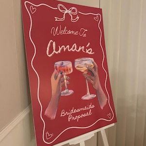 Puede incluir: Un cartel rojo con letras blancas que dice "Welcome to Aman's Bridesmaids Proposal". El cartel está decorado con un lazo blanco y corazones. Dos manos sostienen copas de vino.