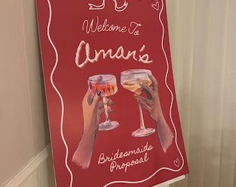 Könnte beinhalten: Ein rotes Schild mit weißen Buchstaben, auf dem "Willkommen zu Aman's Bridesmaids Proposal" steht. Das Schild ist mit einer weißen Schleife und Herzen verziert. Zwei Hände halten Weingläser hoch.