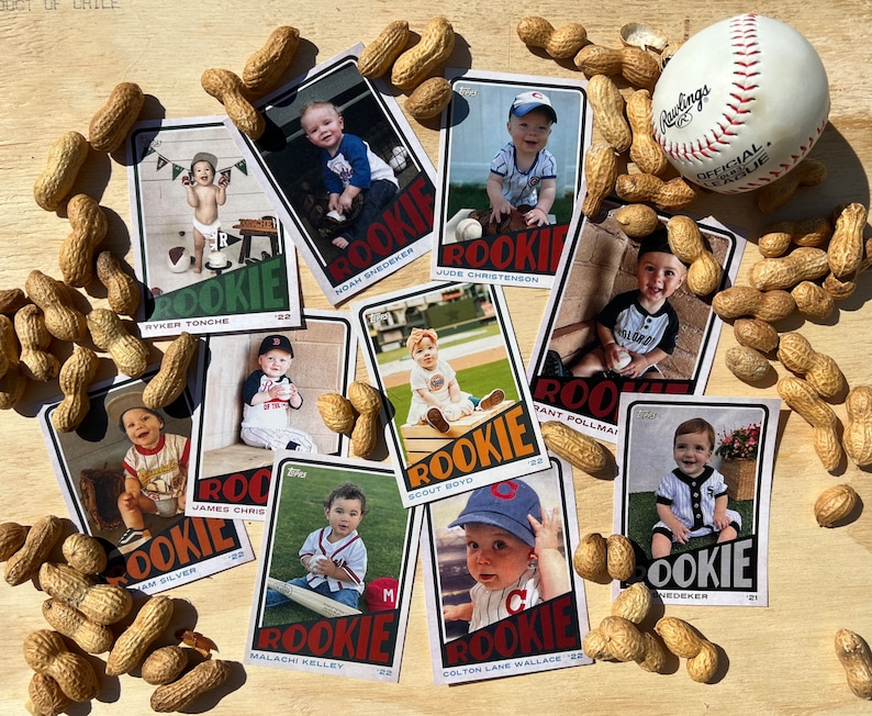 Peut inclure: Plusieurs cartes de baseball de recrues pour b&eacute;b&eacute;s, dispos&eacute;es sur une surface en bois avec des cacahu&egrave;tes et une balle de baseball. Chaque carte pr&eacute;sente une photo de b&eacute;b&eacute; et le mot "ROOKIE".