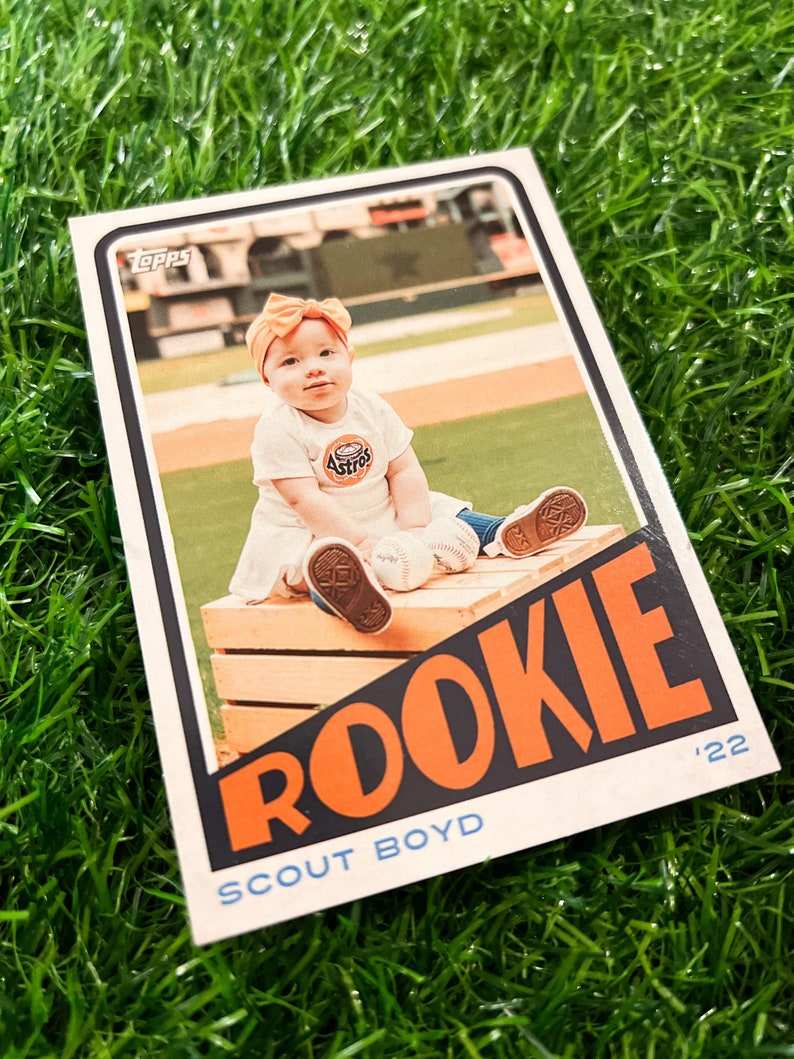 Peut inclure: Carte de baseball avec un b&eacute;b&eacute; portant un t-shirt Astros et un n&oelig;ud. La carte indique "ROOKIE" en orange et "SCOUT BOYD" en bleu. L'arri&egrave;re-plan montre un terrain de baseball.