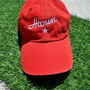 May include: A burnt orange baseball cap with a curved brim. The cap features the word "Htown" embroidered in white script, with a white star below the word. The cap is made of a soft fabric.
