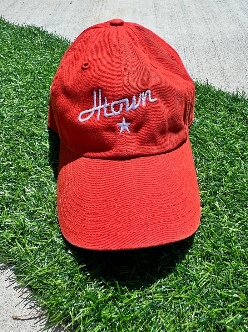 May include: A burnt orange baseball cap with a curved brim. The cap features the word "Htown" embroidered in white script, with a white star below the word. The cap is made of a soft fabric.
