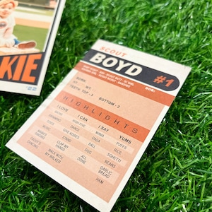 Peut inclure: Deux cartes de baseball vintage sur une surface verte. Une carte pr&eacute;sente un jeune enfant en uniforme de baseball avec le mot "ROOKIE" et le num&eacute;ro "22". L'autre carte est une carte "SCOUT BOYD #1" avec des points forts comme "I LOVE BATHS" et "YUMS PUFFS".