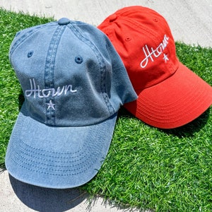 May include: Two baseball caps, one denim blue and one red, rest on artificial green grass. Both caps feature the white embroidered word "Htown" with a star. The caps have a curved brim and a top button.