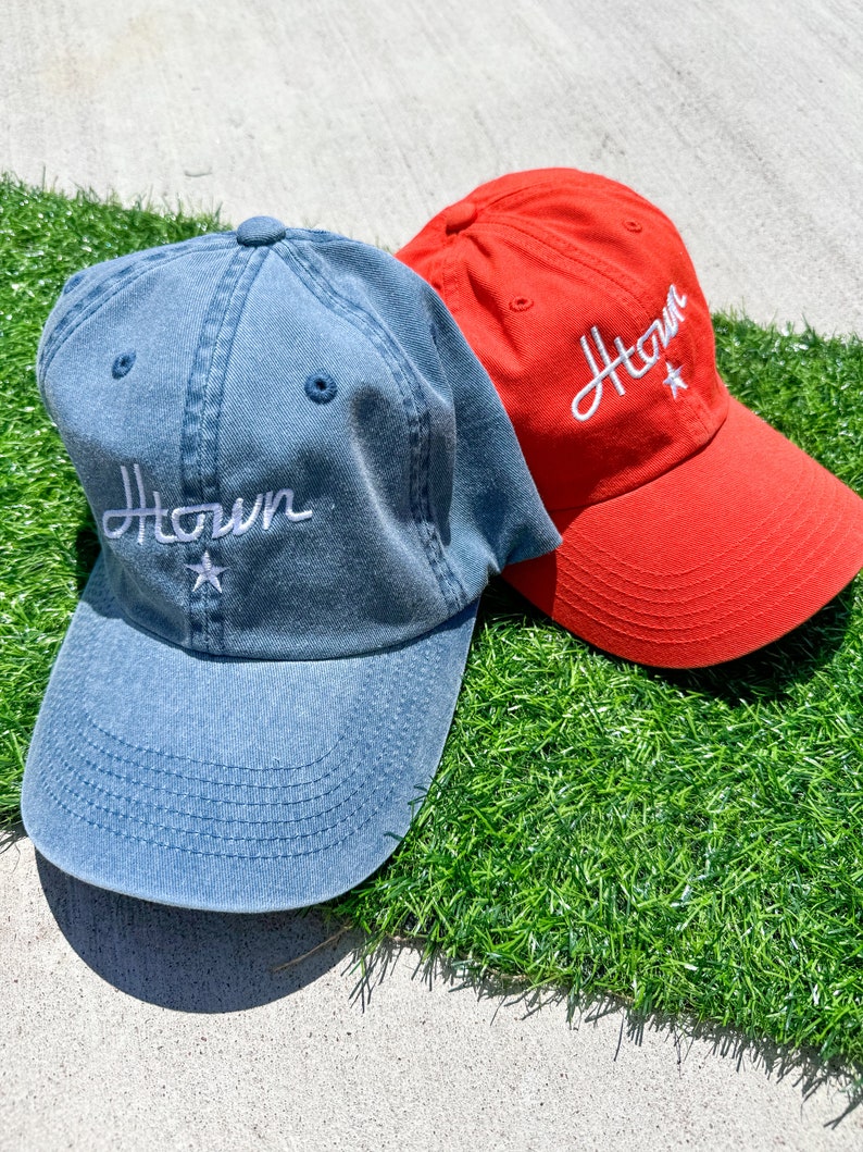 May include: Two baseball caps, one denim blue and one red, rest on artificial green grass. Both caps feature the white embroidered word "Htown" with a star. The caps have a curved brim and a top button.