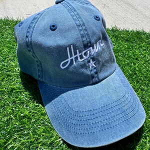 May include: A denim blue baseball cap with the word "Htown" embroidered in white script. A white star is embroidered below the word. The cap is sitting on a bed of green grass.